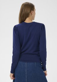Pull-over bleu marine avec poignets et ourlet côtelés, manches bouffantes et encolure ronde, porté avec une jupe en denim.