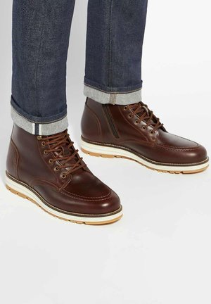 Brown leather ankle boots with a smooth finish, white cushioned sole, and classic lacing, paired with rolled dark denim jeans.