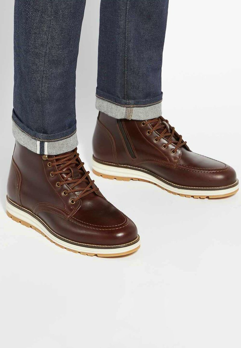 Brown leather ankle boots with a smooth finish, white cushioned sole, and classic lacing, paired with rolled dark denim jeans.