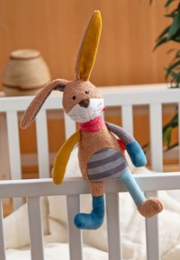 Plush bunny toy with brown, yellow, blue, and gray patchwork hanging on white crib railing in a softly lit nursery.