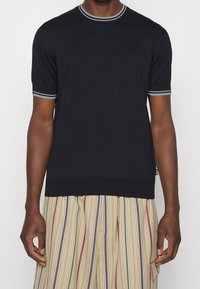 Černý pletený svetr s krátkým rukávem, šedě pruhovaný žebrovaný límec a manžety, v kombinaci s vertikálně pruhovanými kalhotami v béžové, zelené a červené barvě.