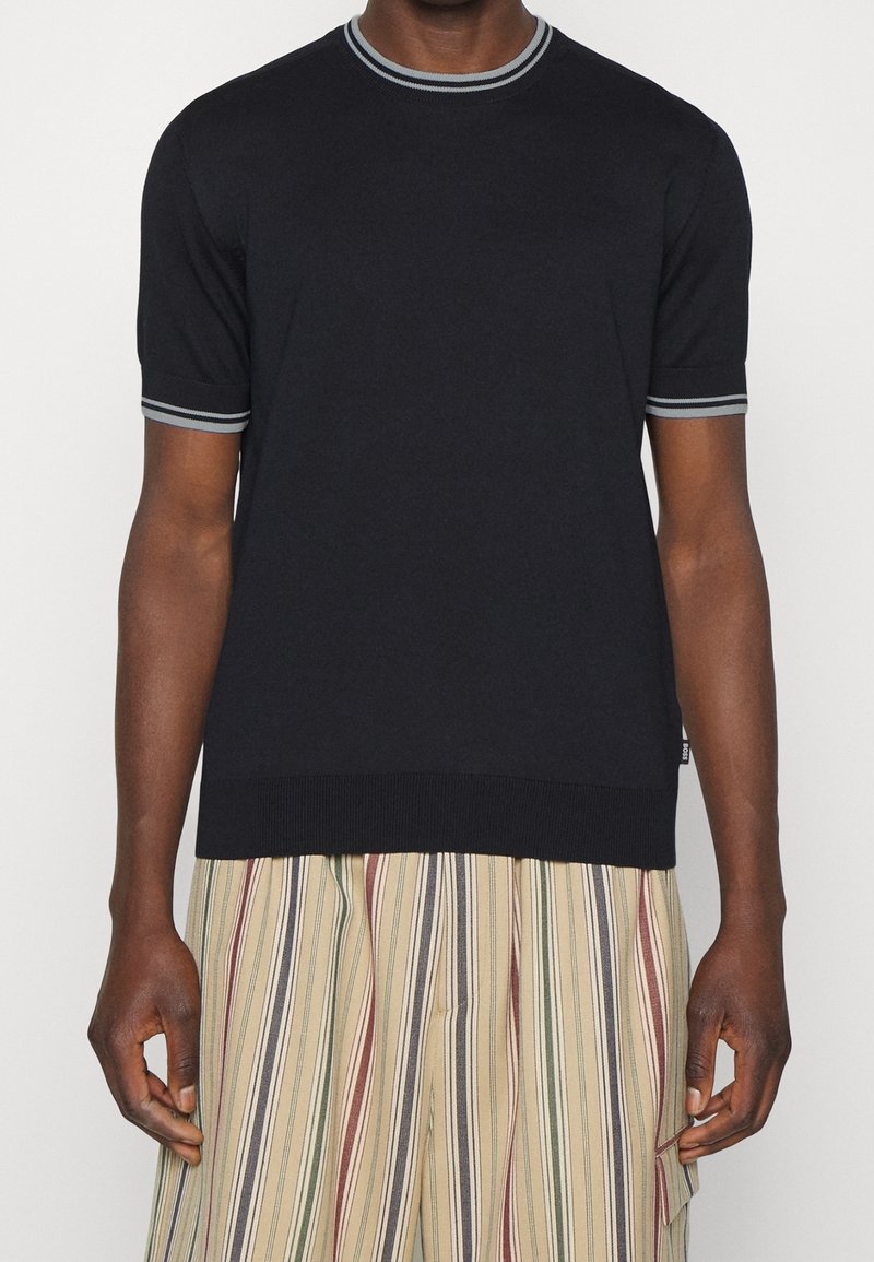 Černý pletený svetr s krátkým rukávem, šedě pruhovaný žebrovaný límec a manžety, v kombinaci s vertikálně pruhovanými kalhotami v béžové, zelené a červené barvě.