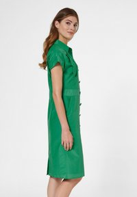 Grünes Kleid mit kurzen Ärmeln, Knopfleiste und seitlichen Schlitzen. Glatte Textur und taillierte Gestaltung. Leichter Stoff.