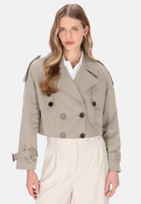 Kurzer Trenchcoat aus hellbeigem Stoff; doppelreihiges Design mit dunklen Knöpfen, weitem Kragen und verstellbaren Manschetten.