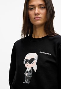 Hanorac negru cu o ilustrație de desen animat a unei figuri cu ochelari de soare, cu textul „KARL LAGERFELD” alb deasupra designului.