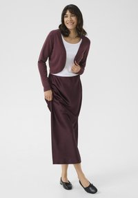 Vínová cardigan přes bílý top, spárovaná s dlouhou vínovou sukní. Černé ploché boty dokončují outfit. Hladká látka, přiléhavý střih.