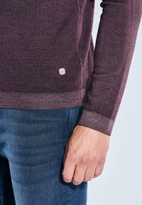 Bordeaux gestrickter Pullover mit gerippten Ärmelbündchen, seitlichem Logo-Tag und glatter Textur, kombiniert mit Jeans. Nahaufnahme von Hand und Ärmeldetail.