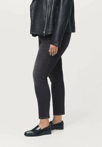 Mujer vestida con chaqueta de cuero negra, jeans ajustados oscuros y mocasines negros, de pie contra un fondo claro y liso.