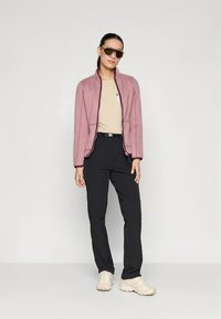 Svetloružová bunda na zips s kontrastnými švami, béžové tričko, čierne nohavice a svetlé tenisky, doplnené o veľké slnečné okuliare.