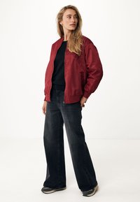 Veste bomber en satin rouge avec des poignets côtelés, associée à un jean noir à jambes larges et des chaussures de sport grises. Cheveux lâchés, posant devant un fond neutre.