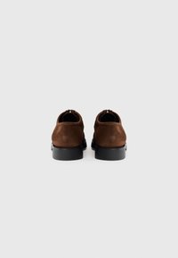 Chaussures en daim marron avec un bout arrondi et des semelles en caoutchouc noir lisses. Présente une couture discrète le long des bords. Vue arrière montrée.
