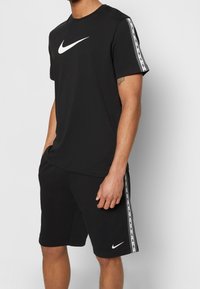 Černé tričko Nike s bílým logem na přední straně a kontrastními bočními pruhy; v kombinaci s černými šortkami, které také mají bílé logo.