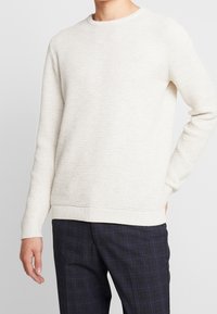 Pull en maille beige clair avec un motif texturé, col rond, et finitions côtelées aux poignets et à l'ourlet, associé à un pantalon à carreaux foncés.