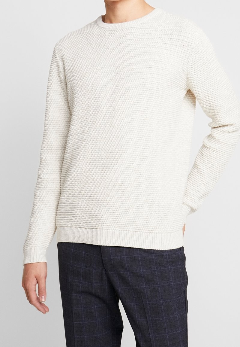 Pull en maille beige clair avec un motif texturé, col rond, et finitions côtelées aux poignets et à l'ourlet, associé à un pantalon à carreaux foncés.
