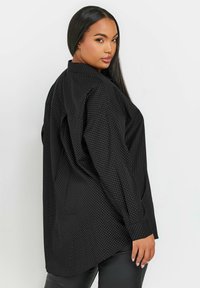 Camicia nera a maniche lunghe con piccoli pois bianchi, design strutturato, polsini con bottone e un retro leggermente allungato. Tessuto morbido.