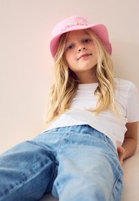 Junges Mädchen mit langen blonden Haaren, trägt einen rosa Hut, ein weißes T-Shirt und blaue Jeans, sitzt entspannt an eine hellfarbige Wand gelehnt.