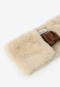 Accessoire en fausse fourrure beige doux avec une texture pelucheuse ; présente une section élastique marron pour l'ajustement ; étiquette visible à l'intérieur.