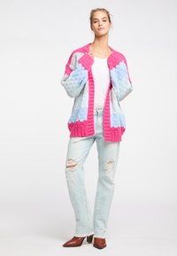 Färgglad stickad kofta med rosa, blå och grå ränder, med en strukturerad stickning och öppen front. Bärs med slitna ljusa jeans.
