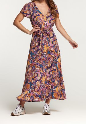 Femme portant une robe midi colorée à motifs, manches courtes avec ceinture à nouer, associée à des baskets blanches de sport, debout sur un fond neutre.