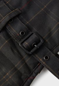 Prim-plan al unei curele din țesătură tartan neagră, cu ochiuri metalice și o cataramă dreptunghiulară mată, neagră, gravată cu "Barbour."