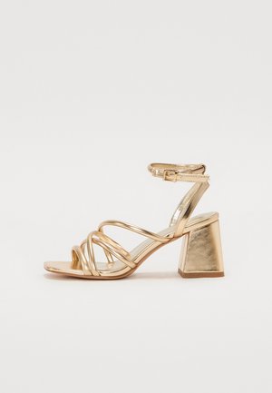 Sandali con tacco oro metallici caratterizzati da un design a strisce, tacco quadrato e cinturino regolabile alla caviglia con fibbia. Texture liscia.