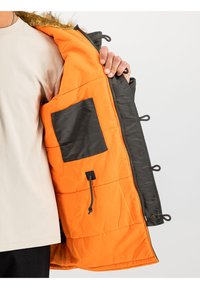 Chaqueta acolchada con capucha forrada de piel, exterior negro y forro naranja brillante. Incluye un bolsillo interior, cremallera y cordón ajustable.