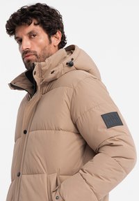 Chaqueta de plumas beige con cremallera frontal, botones a presión y un parche con logo en la manga. Capucha con cordones ajustables y textura suave.