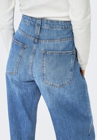 Blå jeans med hög midja, gjorda av denim, med en avslappnad passform, två bakfickor och synliga sömmar runt midjebandet och fickorna.