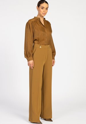 Vrouw in een bruine blouse met knopen en pofmouwen, gecombineerd met hooggewaagde, brede mosterdgele pantalon tegen een effen achtergrond.