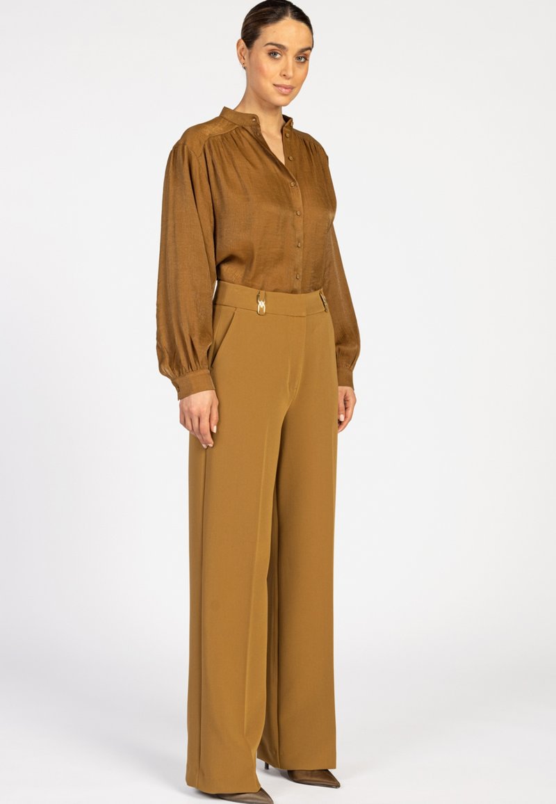 Vrouw in een bruine blouse met knopen en pofmouwen, gecombineerd met hooggewaagde, brede mosterdgele pantalon tegen een effen achtergrond.