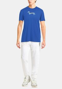 T-shirt in cotone blu con grafica di un bassotto azzurro chiaro, abbinato a pantaloni bianchi e sneaker argentati. Design semplice e casual.