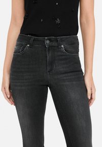 Vaqueros de denim negro con un acabado ligeramente desgastado. Las características incluyen bolsillos frontales, pasadores para el cinturón y una cremallera con cierre de botón.