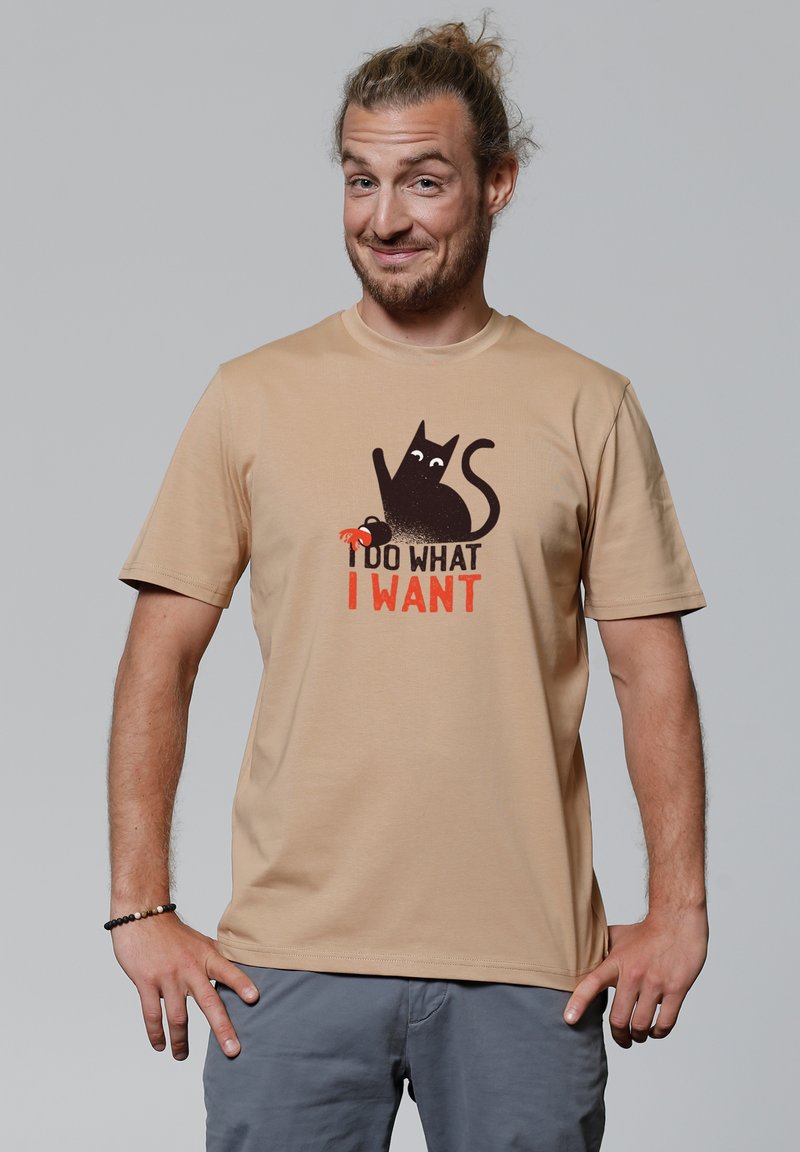 Beige Baumwoll-T-Shirt mit einem schwarzen Katzenmotiv und dem Text "ICH MACH, WAS ICH WILL" in fetter orangefarbener Schrift. Klassisches Rundhalsschnitt-Design.