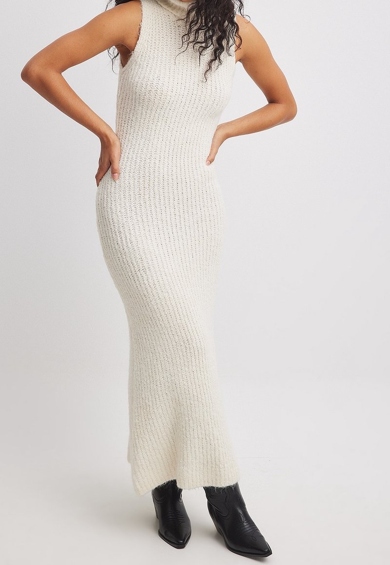 Femme portant une robe maxi en maille crème sans manches avec un col montant et des bottines noires pointues, debout les mains sur les hanches.