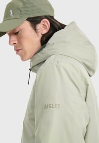 Hellgrüne wasserdichte Jacke mit Kapuze, versehen mit AIGLE-Stickerei, strukturiertem Kragen und belüfteten Akzenten auf der Mütze.