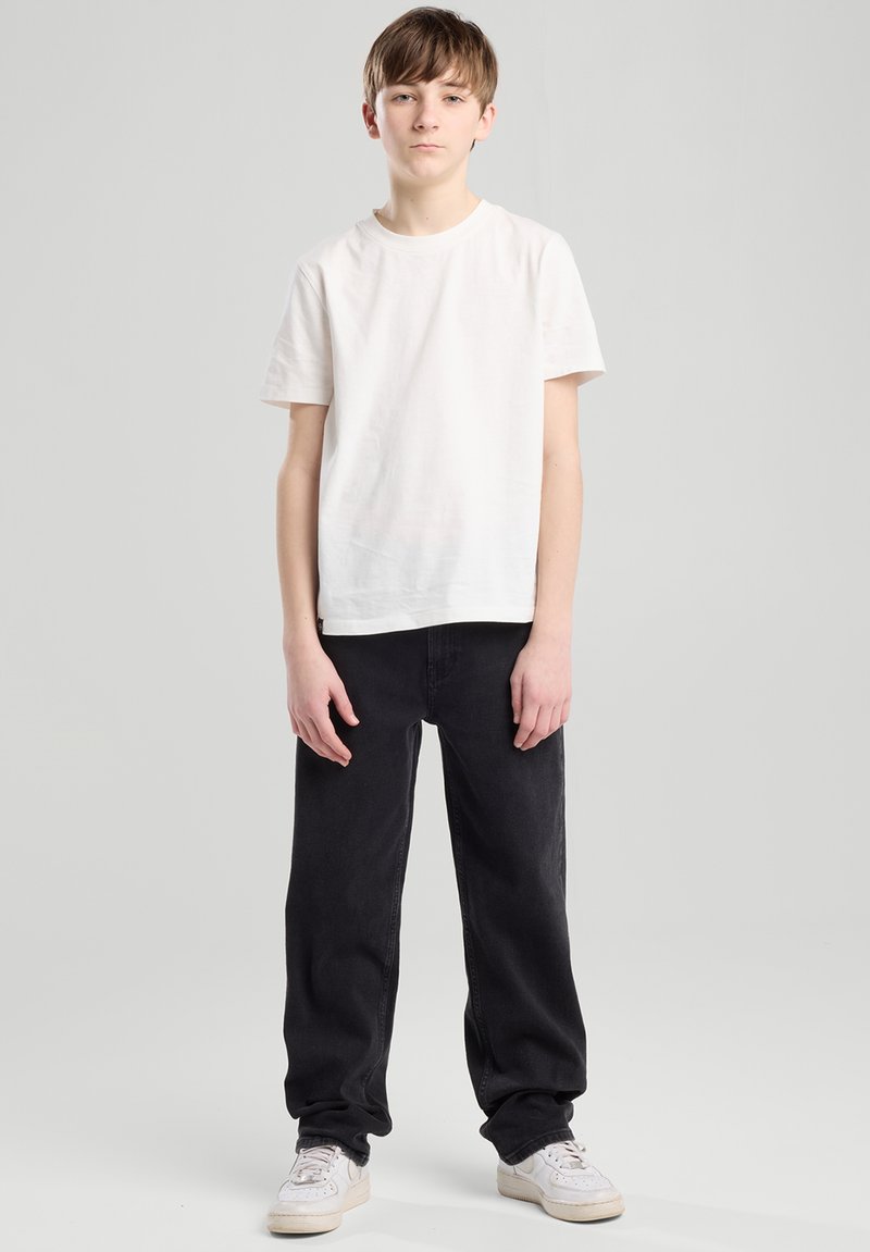 Ein Jugendlicher trägt ein schlichtes weißes Baumwoll-T-Shirt und lockere schwarze Jeans, steht vor einem neutralen Hintergrund und trägt weiße Sneakers.