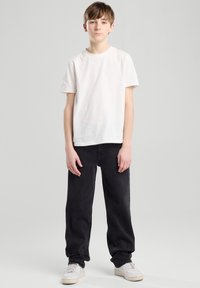 Nuori, joka on pukeutunut yksinkertaiseen valkoiseen puuvillapaitaan ja löysiin mustiin farkkuihin, seisoo neutraalia taustaa vasten valkoisissa lenkkareissa.