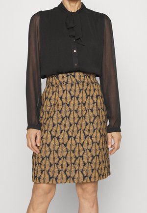 Femme portant un chemisier noir transparent à manches longues et une jupe genou avec des motifs de feuilles marron sur un fond sombre.