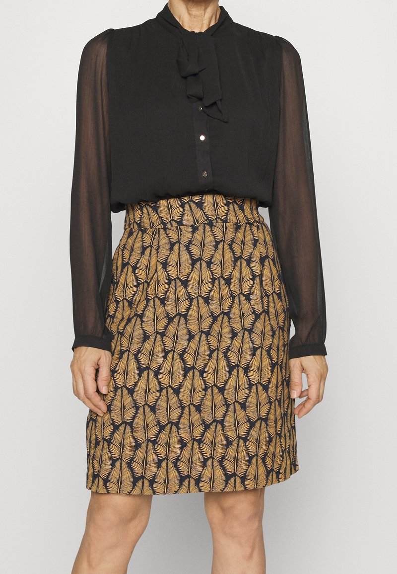 Femme portant un chemisier noir transparent à manches longues et une jupe genou avec des motifs de feuilles marron sur un fond sombre.