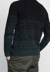 Pull en maille vert foncé et noir avec un motif en dégradé, poignets et ourlet côtelés, surface texturée et coupe ajustée.