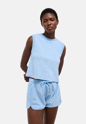 Femme portant un haut sans manches en éponge bleu clair et un short assorti, debout avec les bras dans le dos devant un fond uni.