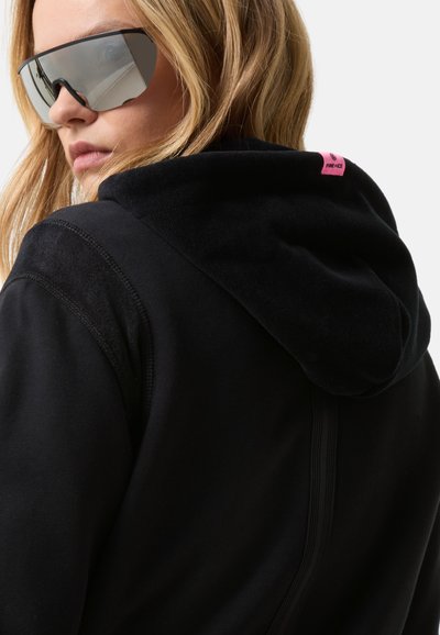 Blondýna v nočních brýlích s velkými reflektivními skly a černé mikině s malou růžovou nášivkou na zadní straně kapuce.