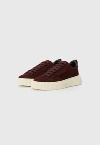 Une paire de baskets en daim marron foncé avec des semelles en caoutchouc ribbées épaisses de couleur blanc cassé et des lacets assortis marron sur un fond clair uni.