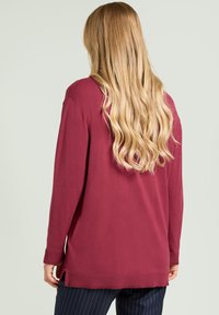 Bordowy sweter z długim rękawem o luźnym kroju, z prążkowanymi mankietami, bocznymi rozcięciami i teksturowanym wykończeniem, noszony na spodniach w prążki.