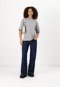 Šedé oversized tričko, tmavě modré zvonové džíny a černé platformové boty. Modelka nese světle šedou tašku přes rameno. Hladké textury a ležérní design.
