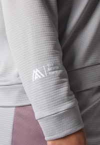 Top grigio a maniche lunghe con un motivo testurizzato, caratterizzato da un logo bianco sulla manica. Il tessuto appare liscio con un design aderente.