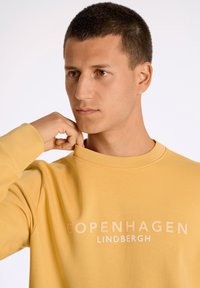 Gul sweatshirt med rund halsringning, med prägling av texten "COPENHAGEN" och "LINDBERGH" i en kontrasterande färg, slät tygstruktur.
