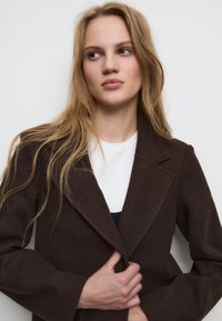 Bruine wolmix jas met een geknipt revers, lange mouwen en een gestructureerd oppervlak. Model draagt een witte top eronder.