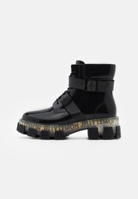 Bottines en cuir verni noir avec une semelle épaisse et transparente ornée d'un logo jaune, maintenues par une large sangle noire et des lacets.
