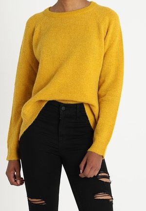Personne portant un pull à manches longues jaune moutarde et un pantalon noir taille haute avec les genoux déchirés sur un fond blanc.